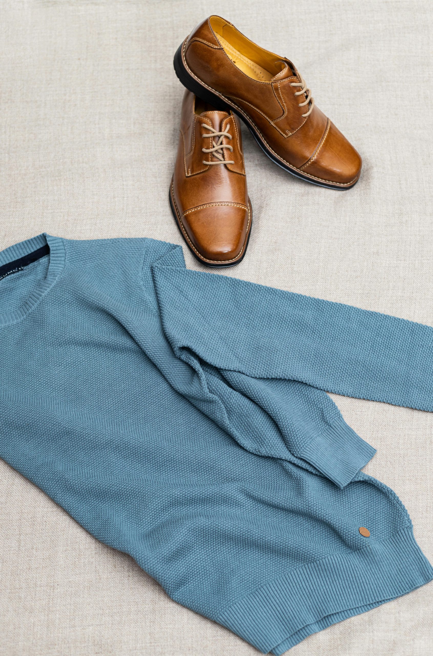 Brown leather shoes paired with a blue sweater on a beige textile backdrop, showcasing men's fashion.